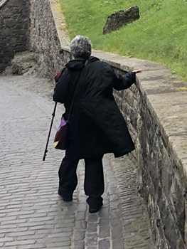 Susan with cane on cobblestones using wall for balancing