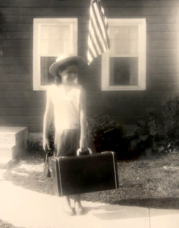 little girl carrying suitcase: in striped dress in front of stipes of flag